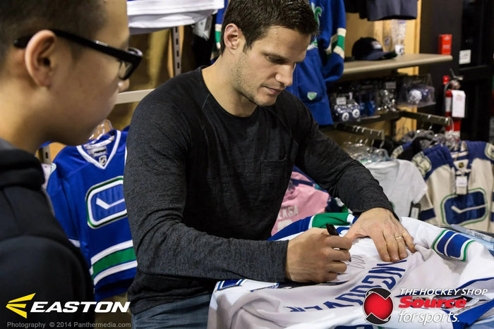 Hockey player signing a Vancouver Canucks jersey at The Hockey Shop, surrounded by hockey gear.