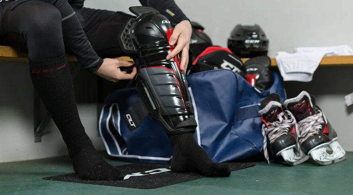 Hockey player putting on CCM shin guards in locker room with equipment bag, helmets, and skates