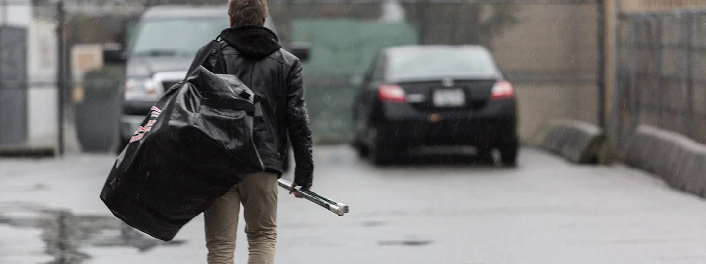 Hockey player carrying large hockey equipment bag and sticks in rainy parking lot