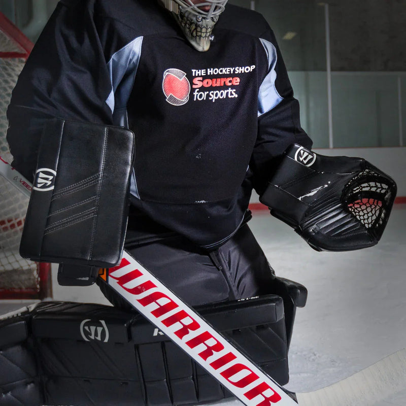 Hockey goalie in black Warrior gear with trapper, blocker, and stick on ice rink, The Hockey Shop jersey.