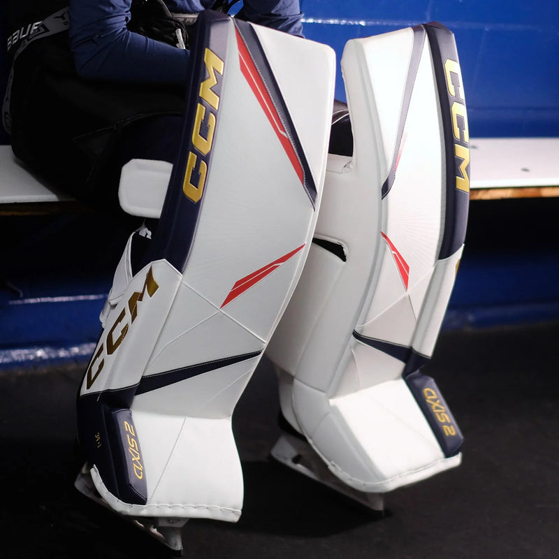 CCM Axis 2 goalie leg pads in white, navy, and gold at hockey rink, The Hockey Shop