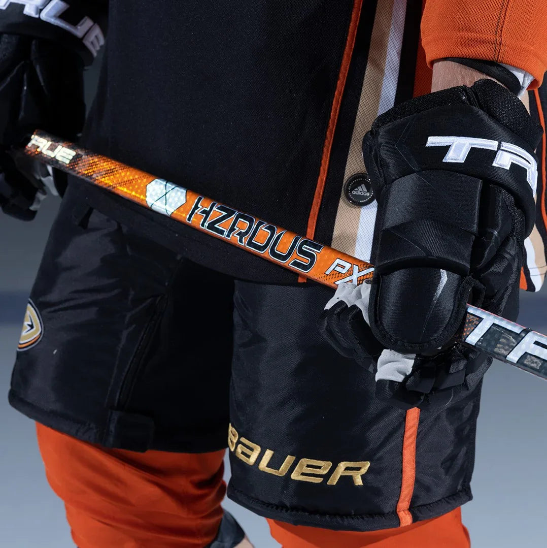 Close-up of a hockey player in Bauer shorts, orange pants, True gloves, holding HZRDUS PX stick