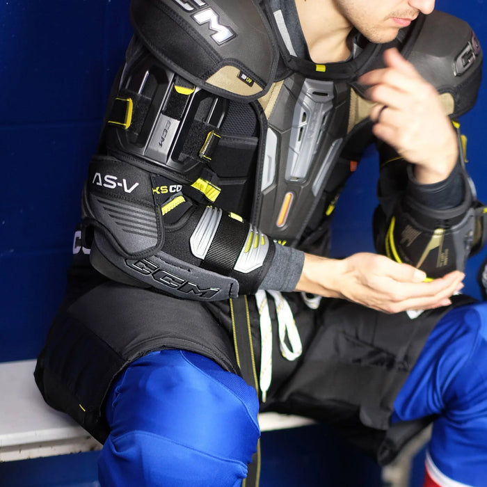 Hockey player wearing CCM AS-V protective gear and blue pants on locker room bench at The Hockey Shop