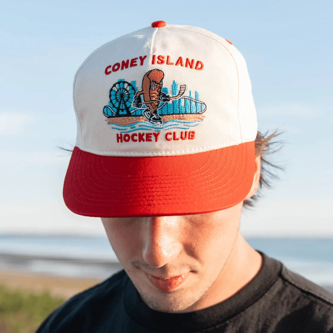 Person wearing a Coney Island Hockey Club cap with a scenic design against a clear sky.